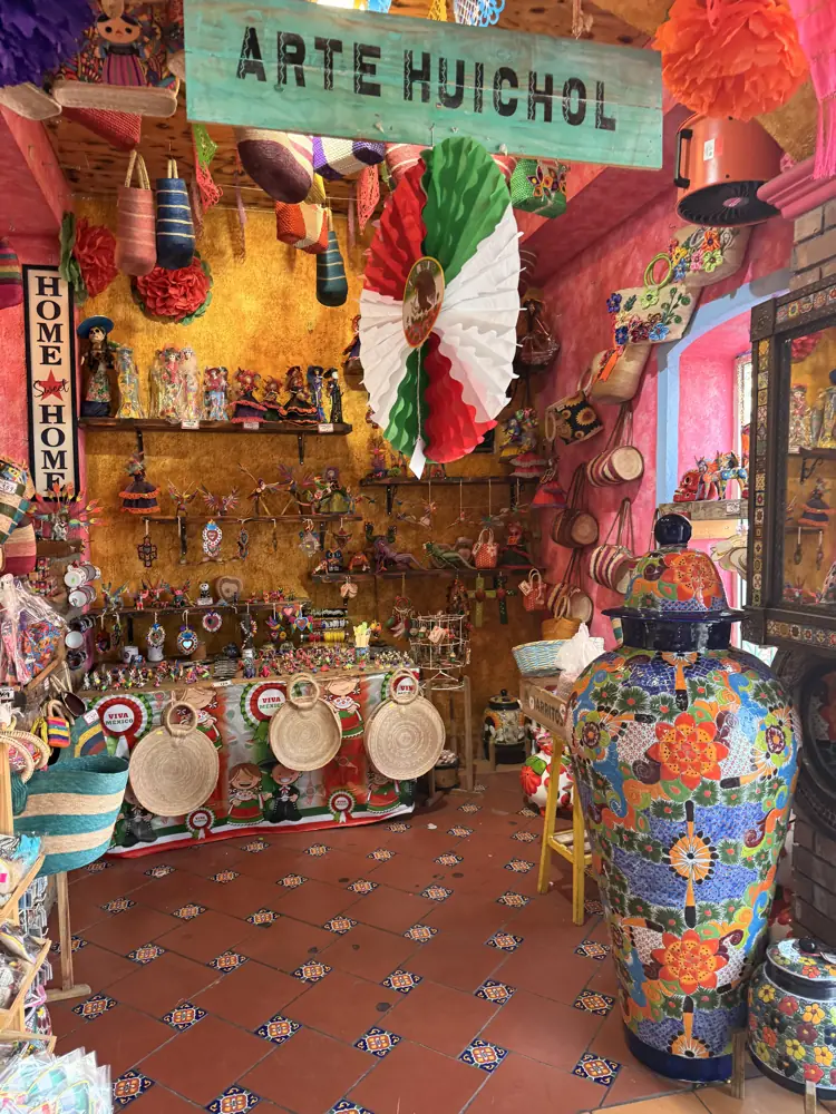 Sala rotulada Arte Huichol con jarrón de talavera al frente, sombreros y artesanía colgada en la pared rosa