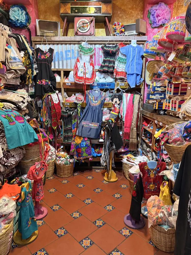 Vestidos y blusas bordadas colgadas sobre piso de mosaico, con figuras de catrina y canastas